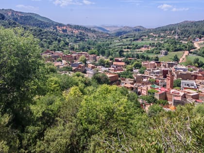 View over the village of Zaouia d’Ifrane, earthen houses and a minaret among trees, the green hills of the Middle Atlas behind