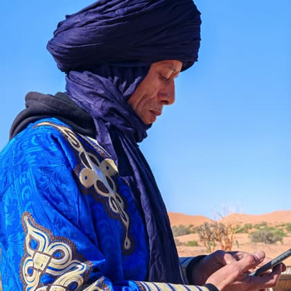 Ali in a blue robe and turban, phone in hand, the dunes behind him