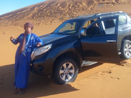 Ali in a blue robe beside the four-wheel drive at the foot of a dune